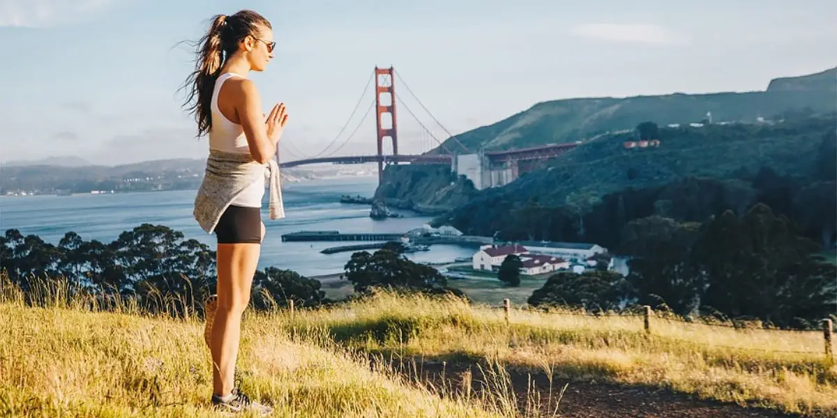 A woman stands on a grassy hill doing a yoga pose with hands pressed together. She is wearing a white tank top, black shorts, sunglasses, and a jacket tied around her waist. In the background, near some of the best wellness hotels North Bay has to offer, is the iconic Golden Gate Bridge and a scenic view of hills and water.