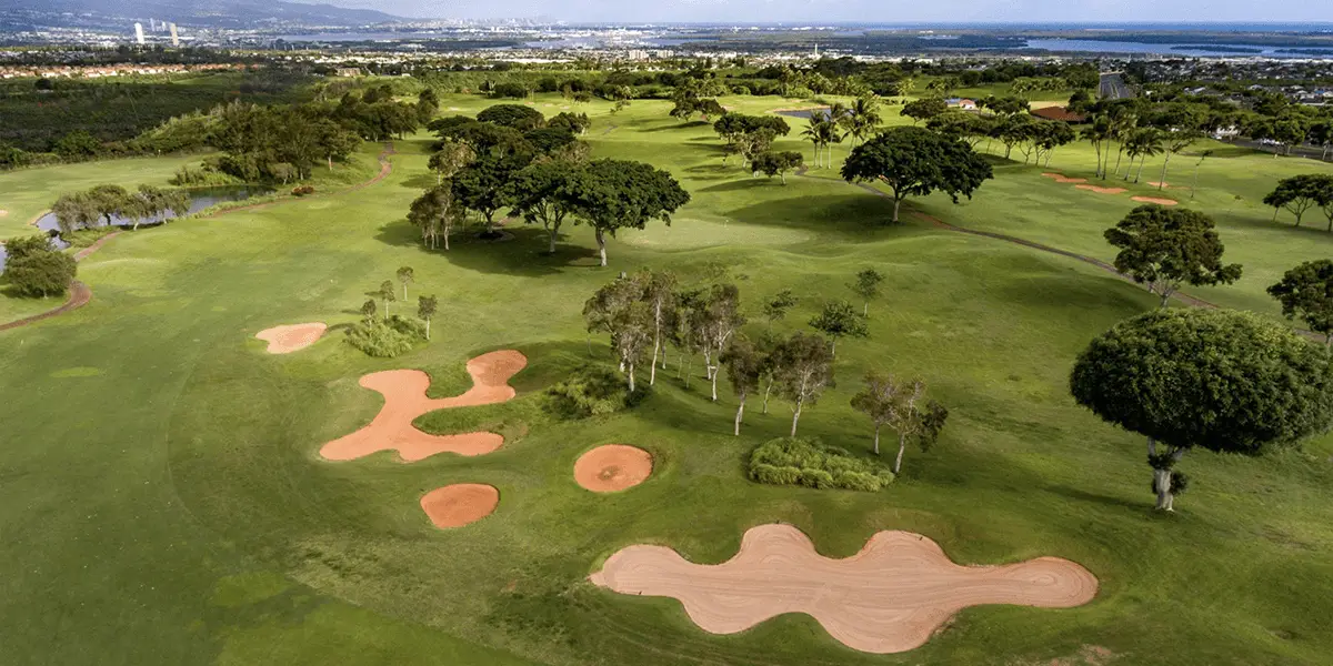 Aerial view of one of the best golf courses in Oahu, featuring lush green fairways, multiple sand bunkers, and scattered trees. The well-manicured landscape extends to a distant city skyline with mountains and a clear blue sky in the background.