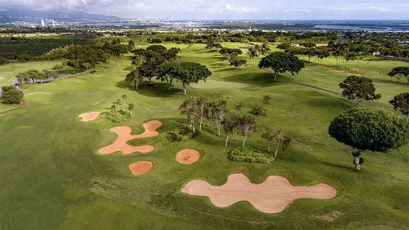 Aerial view of a lush golf course, featuring several sand traps, clusters of trees, and manicured greens. This scenic landscape stretches out with distant city buildings on the horizon and expansive blue skies above. Truly one of the best golf courses Oahu has to offer.