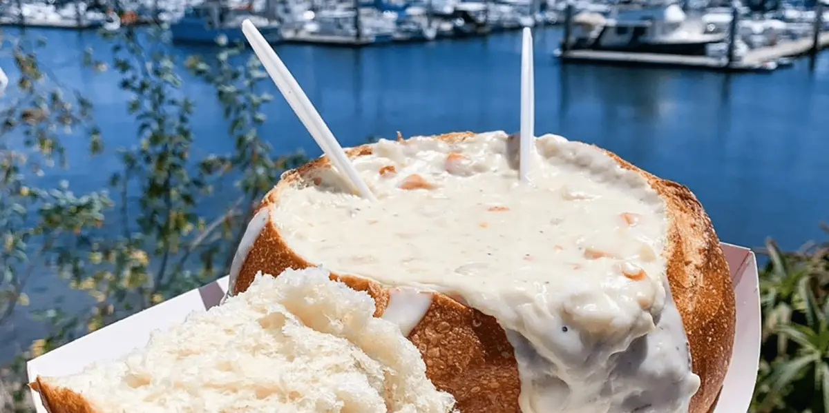 A bread bowl filled with creamy clam chowder, touted as the best on the Monterey Peninsula, is held up against a backdrop of a marina with several docked boats and calm water. Two clear plastic spoons stick out, and a piece of bread from the bowl is placed to the side, ready to be dipped.