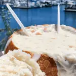 A bread bowl filled with creamy clam chowder, touted as the best on the Monterey Peninsula, is held up against a backdrop of a marina with several docked boats and calm water. Two clear plastic spoons stick out, and a piece of bread from the bowl is placed to the side, ready to be dipped.