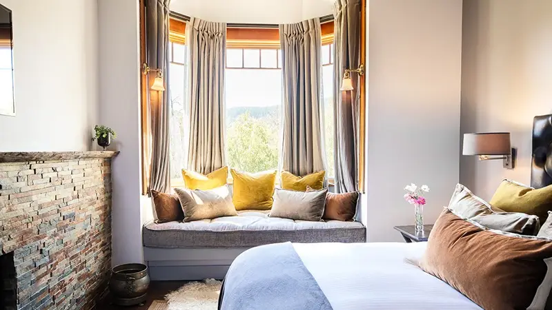 A cozy bedroom reminiscent of the best luxury hotels North Bay has to offer, featuring a large bay window with curtains, a window seat adorned with various throw pillows, and a bed topped with cushy pillows and a brown blanket at the foot. The room bathes in natural light and includes a small side table with flowers in a vase.