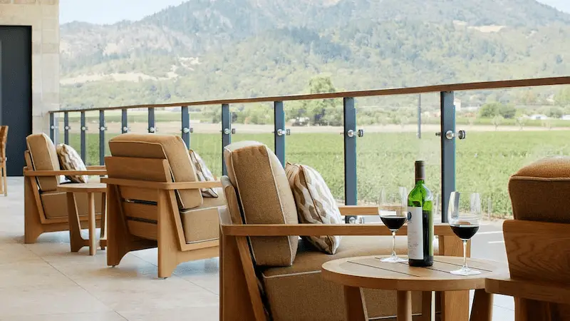 A scenic outdoor patio features cushioned wooden chairs with small tables, each adorned with wine glasses and a bottle. Clear glass railing provides an unobstructed view of green fields and distant mountains under a partly cloudy sky, reminiscent of the best dining wineries in Napa Valley.