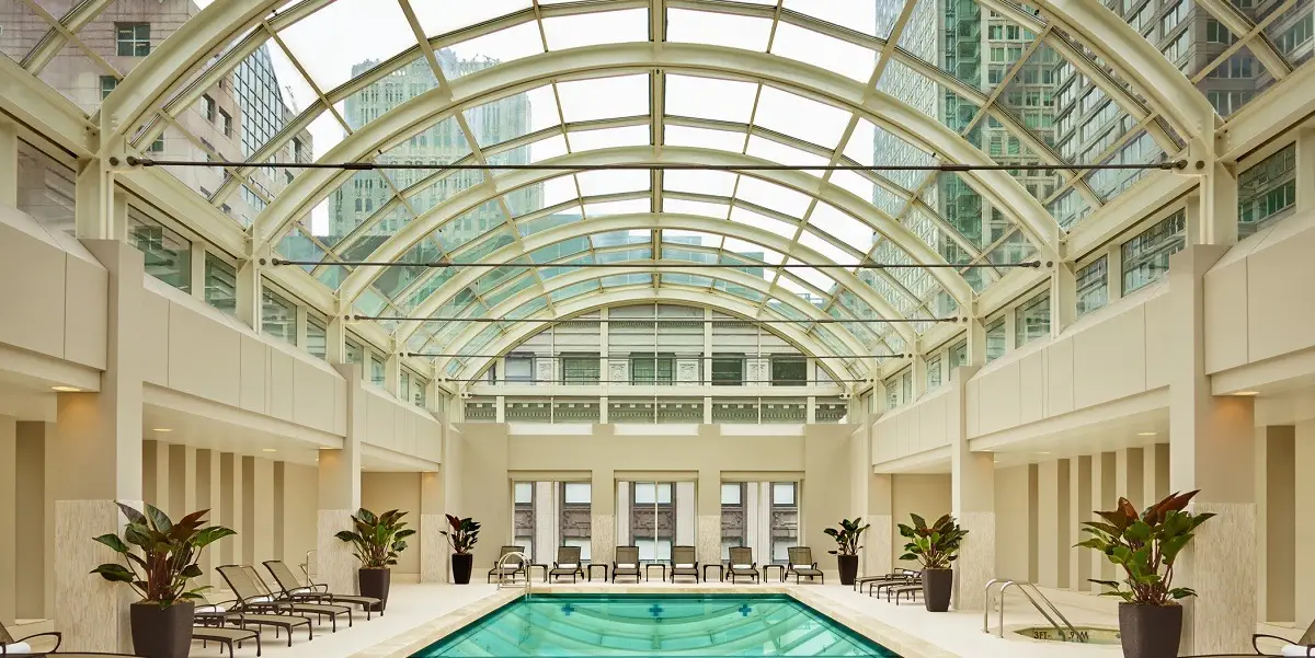 A luxurious indoor swimming pool with clear, blue water is housed under a glass-domed ceiling. Sunlight filters through, illuminating the surrounding lounge chairs and potted plants. Tall buildings can be seen through the large windows and ceiling above, making it a hidden gem among romantic hotels in San Francisco.