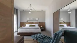 Modern bedroom with minimalist decor, inspired by the simplicity of Northern California Missions. Features a large bed with white linens and a green accent pillow. Wooden headboard and bedside tables with hanging lights. At the foot of the bed are two blue tufted benches. Large mirror reflecting the room's setup.