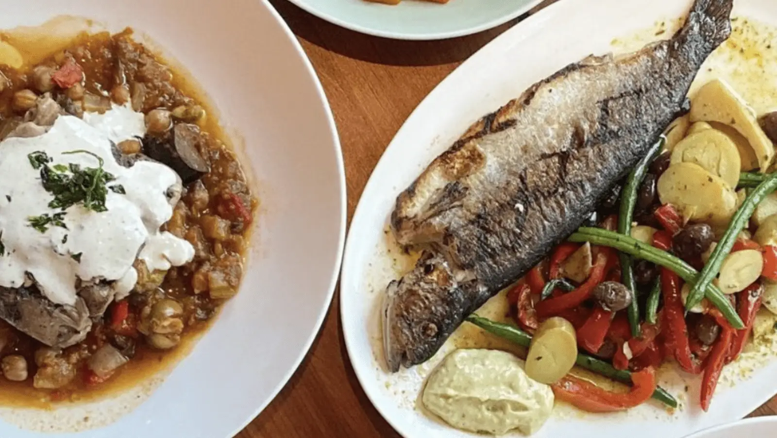 Two plates of food are shown, showcasing the best dinner on the Monterey Peninsula. The left plate has a hearty stew topped with a dollop of creamy sauce and garnished with herbs. The right plate features a whole grilled fish with crispy skin, accompanied by sautéed vegetables, including green beans, peppers, and potatoes.