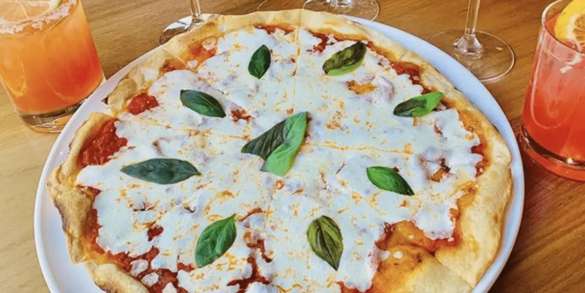 A Margherita pizza topped with melted mozzarella cheese and fresh basil leaves. The pizza is on a white plate, surrounded by glasses of orange and red drinks on a wooden table, making it the best dinner on the Monterey Peninsula.