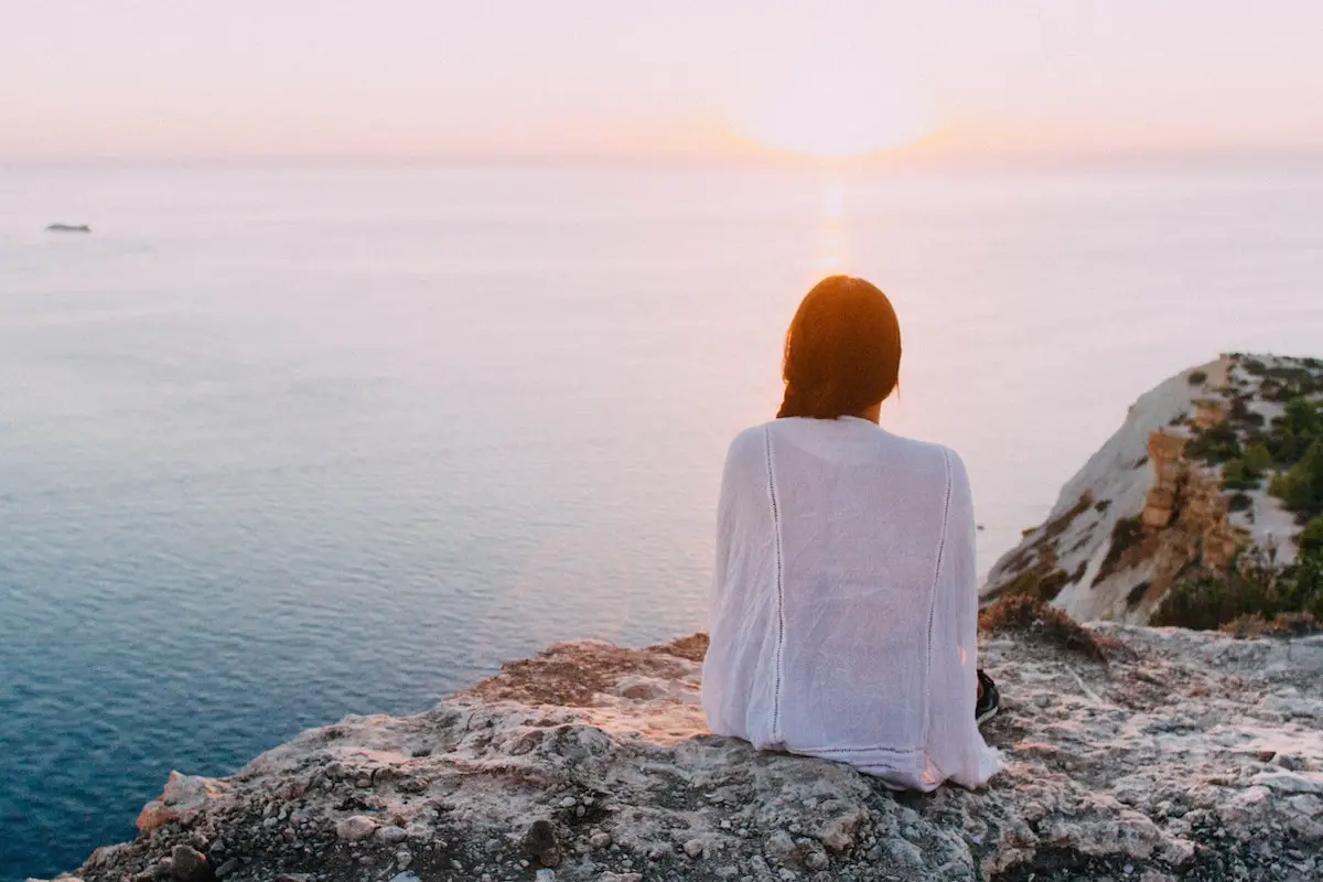 A person with long hair, wearing a white shirt, sits on a rocky cliff overlooking a serene ocean as the sun sets. The sky is a gradient of soft pinks and oranges, reflecting on the calm water below—much like BTC's ever-changing yet mesmerizing charts.