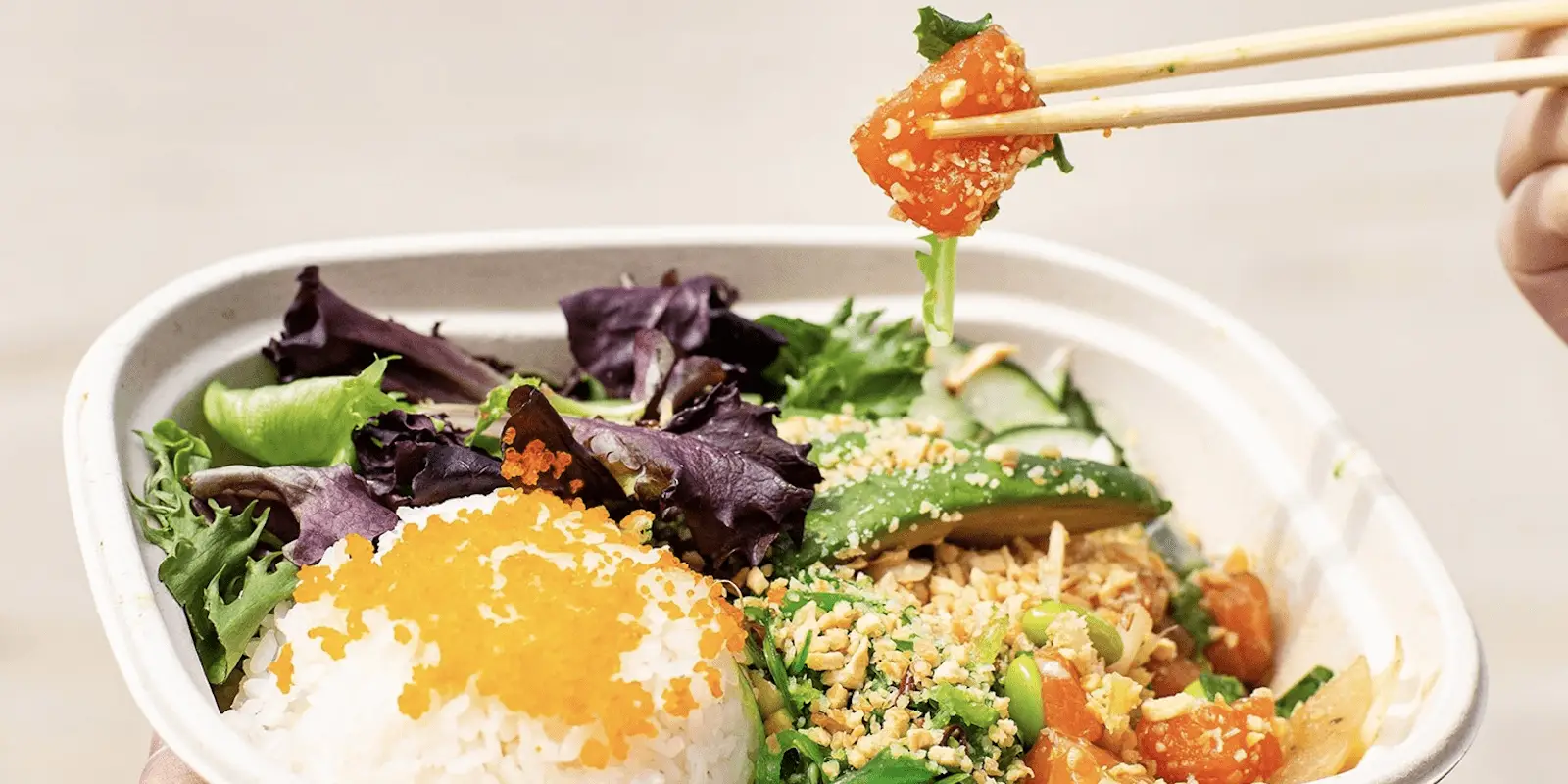 A close-up of a healthy meal in a bowl, featuring a portion of white rice topped with orange fish roe, fresh leafy greens, diced salmon being held with chopsticks, cucumber slices, crushed nuts, and edamame. This dish could easily be found at the best poke spots in Waikiki Oahu.