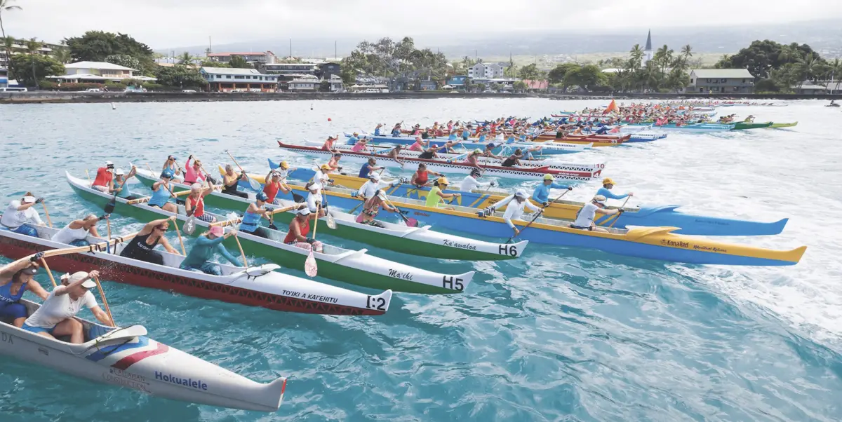 A large group of rowers in colorful outrigger canoes line up at the starting line of a race on a bright, clear day. The canoes and paddlers are clustered closely, creating an energetic and competitive atmosphere on the blue water, reminiscent of traditional Hawaiian holidays. The shoreline is visible in the background.