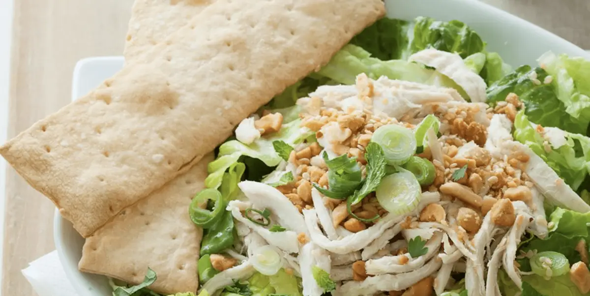 A fresh salad composed of shredded chicken, chopped nuts, and green onions, served on a bed of leafy greens. Two large rectangular crackers are placed on the side in a white bowl, making it one of the best lunch options in North Bay.