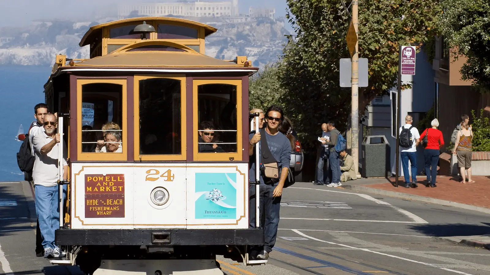 A classic San Francisco cable car labeled "Powell and Market" travels along a street. Several passengers are riding, some seated and others standing, embarking on their San Francisco public transportation adventures. People stroll on the sidewalk in the background, with a partial view of a hilly landscape.