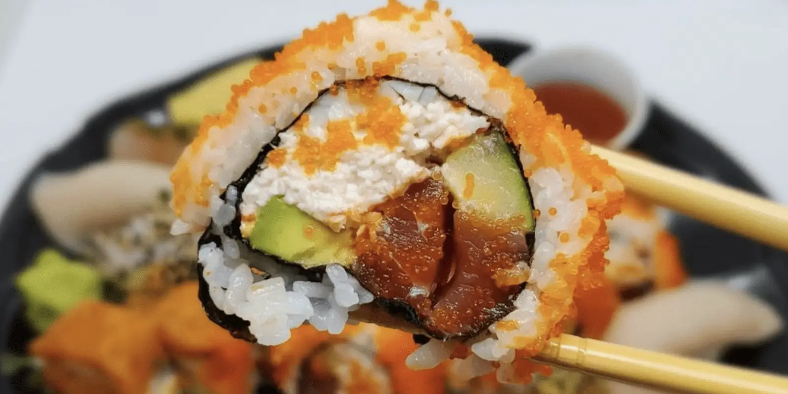 Close-up of a sushi roll held by chopsticks. The roll, from the best sushi on the Big Island, contains rice, avocado, cucumber, and fish, topped with orange fish roe and a creamy white filling. Other rolls and a bowl of soy sauce are blurred in the background.