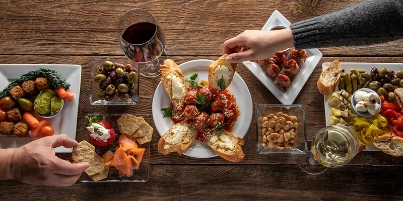A wooden table is filled with a variety of appetizers, including dips, olives, stuffed peppers, bruschetta, and bread. Two people reach for food, and there are glasses of red and white wine on the table. The scene exudes a casual, social dining atmosphere reminiscent of the best wine bars in the Bay Area.