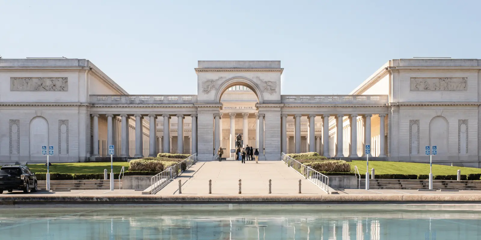 A grand neoclassical building with columns and an arch, flanked by two wings. A wide pathway leads to the entrance, lined with sculptures and greenery. The foreground features a reflective pool. The sky is clear and bright—one of the many stunning sights as you explore San Francisco.