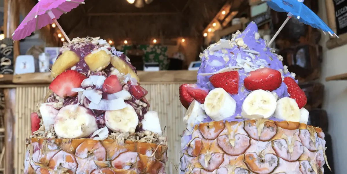 Two hollowed-out pineapples filled with colorful fruit and ice cream, topped with fresh strawberries, bananas, coconut flakes, and granola. Each dessert is garnished with a small paper umbrella. The background shows a cozy, rustic cafe setting known for the best smoothies and acai bowls on Hawaii Island.