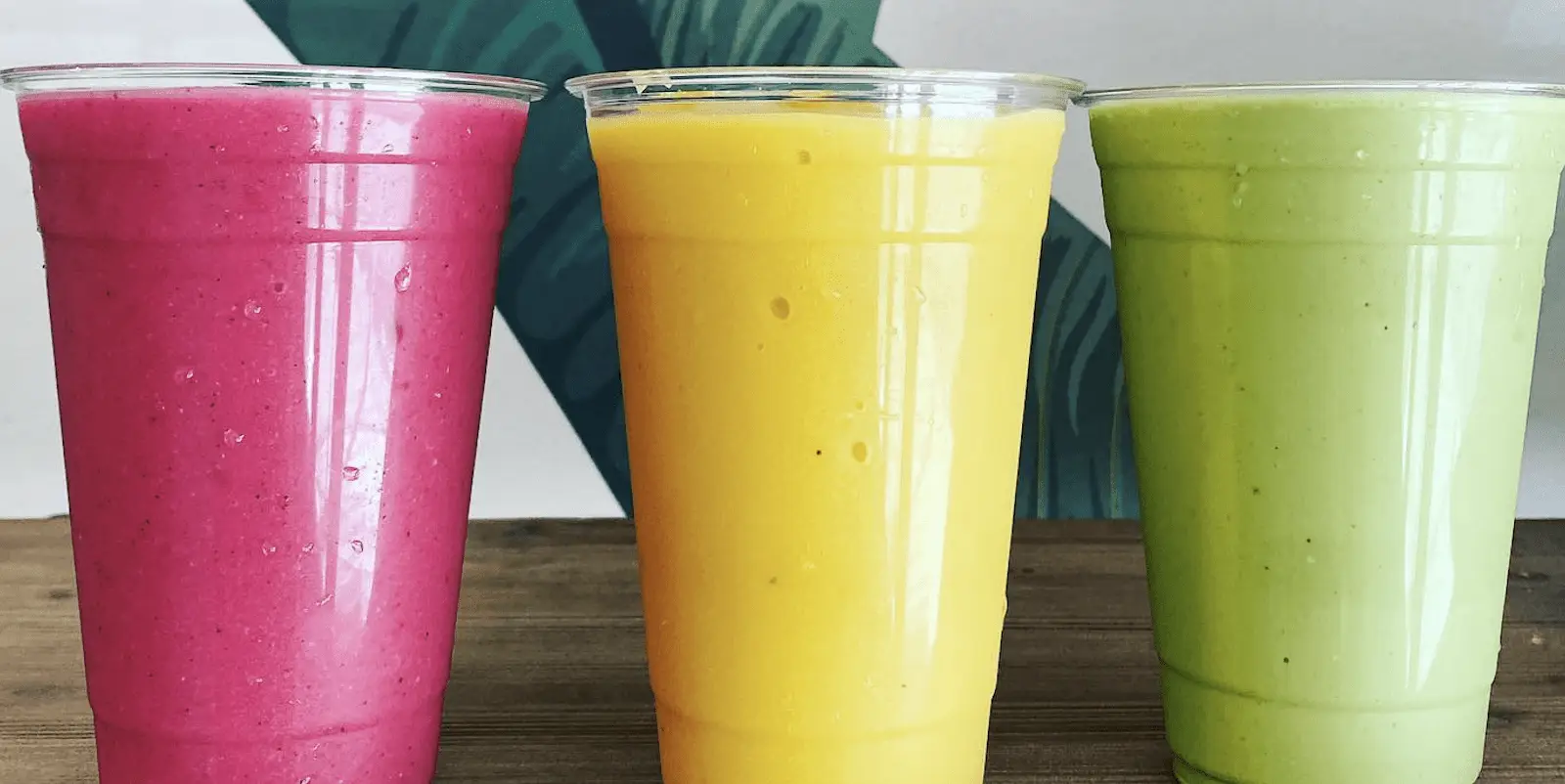 Three plastic cups filled with different colored smoothies are placed in a row on a wooden surface. The left cup has a pink smoothie, the middle cup contains a yellow smoothie, and the right cup is filled with a green smoothie—perfectly reminiscent of the vibrant smoothies and acai bowls from Kauai.