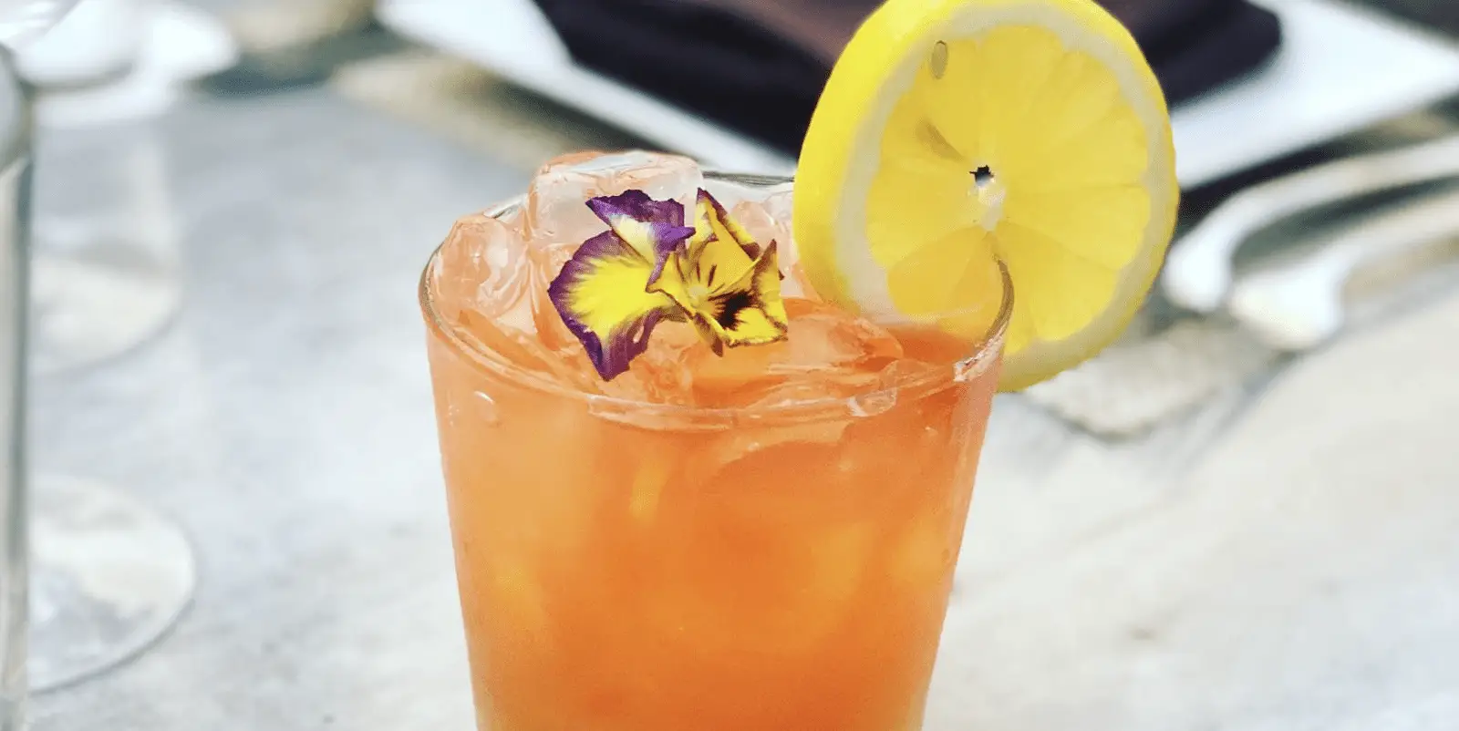 A refreshing orange-hued cocktail served over ice in a clear glass, garnished with a vibrant yellow lemon slice and a delicate purple and yellow flower. The background is blurred and out of focus, suggesting an outdoor or casual setting—perfect for the best happy hour in Kauai.