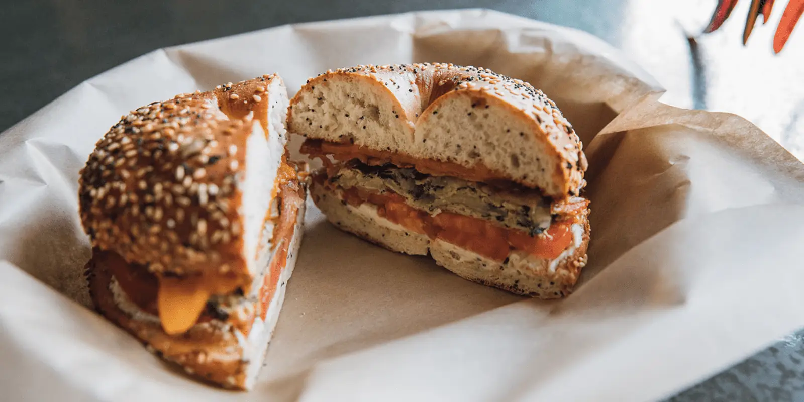 A halved bagel sandwich on parchment paper in a basket. The bagel, topped with sesame and poppy seeds, is filled with cheese, egg, tomato slices, and other ingredients. Neatly cut to reveal its fillings, this could easily be part of the Best Breakfast Kauai has to offer.