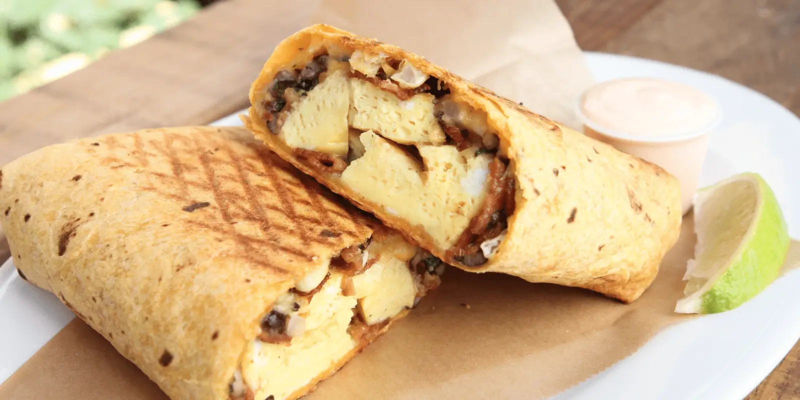 A grilled breakfast burrito, cut in half to reveal scrambled eggs, cheese, and vegetables, is served on brown parchment paper with a slice of lime and a small cup of dipping sauce. Some say it's the best breakfast on the Big Island.