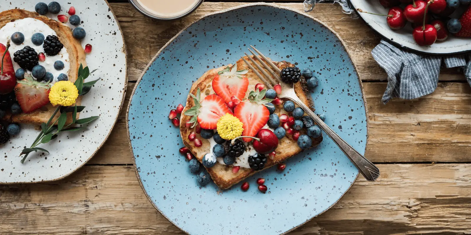 A rustic wooden table holds a blue speckled plate with French toast topped with strawberries, blueberries, blackberries, pomegranate seeds, and a yellow flower. A fork rests on the plate. Nearby, a smaller plate and bowl hold more toast and fresh berries—truly the best breakfast Ko Olina Oahu offers.