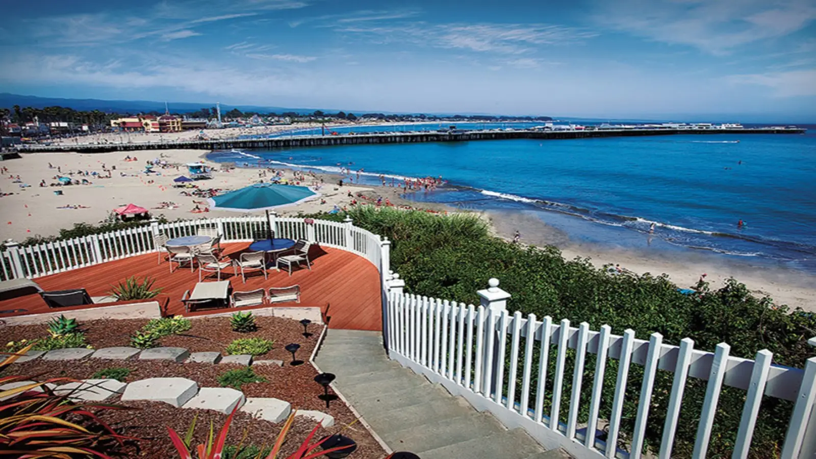A scenic beach view with a vibrant blue ocean. In the foreground, there's a wooden deck with tables and chairs surrounded by a white fence. The sandy beach below has sunbathers and umbrellas, and a long pier stretches out over the water. Nearby are some of the best group hotels South Bay has to offer.