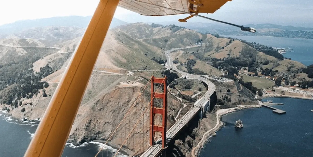 Aerial view from a Seaplane Adventures aircraft showing the Golden Gate Bridge spanning over a bay between two hilly landscapes. The red-orange bridge contrasts with the green hills and the blue waters below, while roads and buildings are visible in the distance.