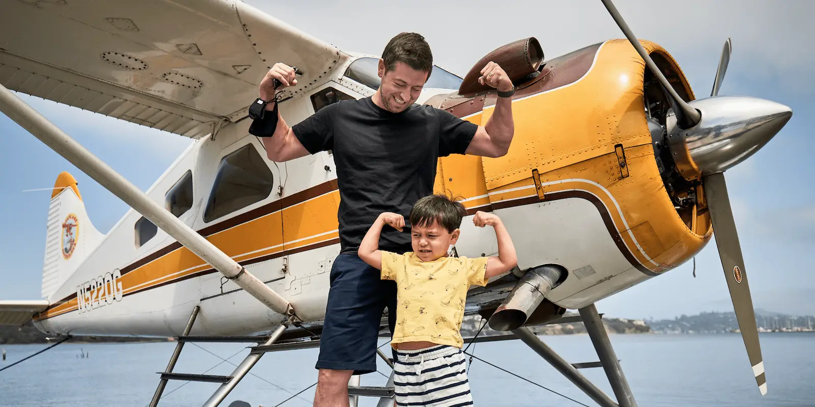 A man and a young boy are standing in front of a yellow and white seaplane, enjoying their summer with kids. Both are flexing their arm muscles and smiling. The man is wearing a black shirt and blue shorts, while the boy is in a light yellow shirt and striped shorts. The sky is clear and blue.