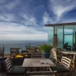 A serene outdoor seating area of one of the best restaurants in Big Sur features tables and chairs, with a modern glass-walled section extending from the main building. A fire pit is surrounded by potted plants, and the open-air deck overlooks a calm ocean under a partly cloudy sky.