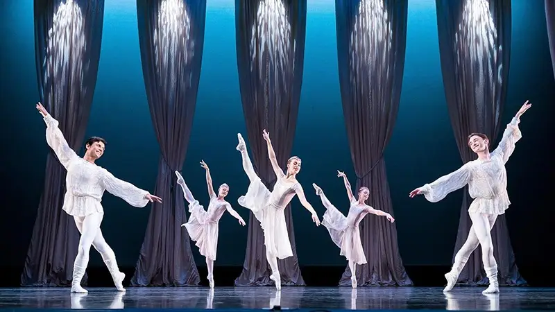 Five ballet dancers are performing on stage. Dressed in white costumes, they strike an elegant pose with one leg extended high. The background features tall, flowing curtains and dramatic lighting, reminiscent of the enchanting performances you can catch during December on the Monterey Peninsula.