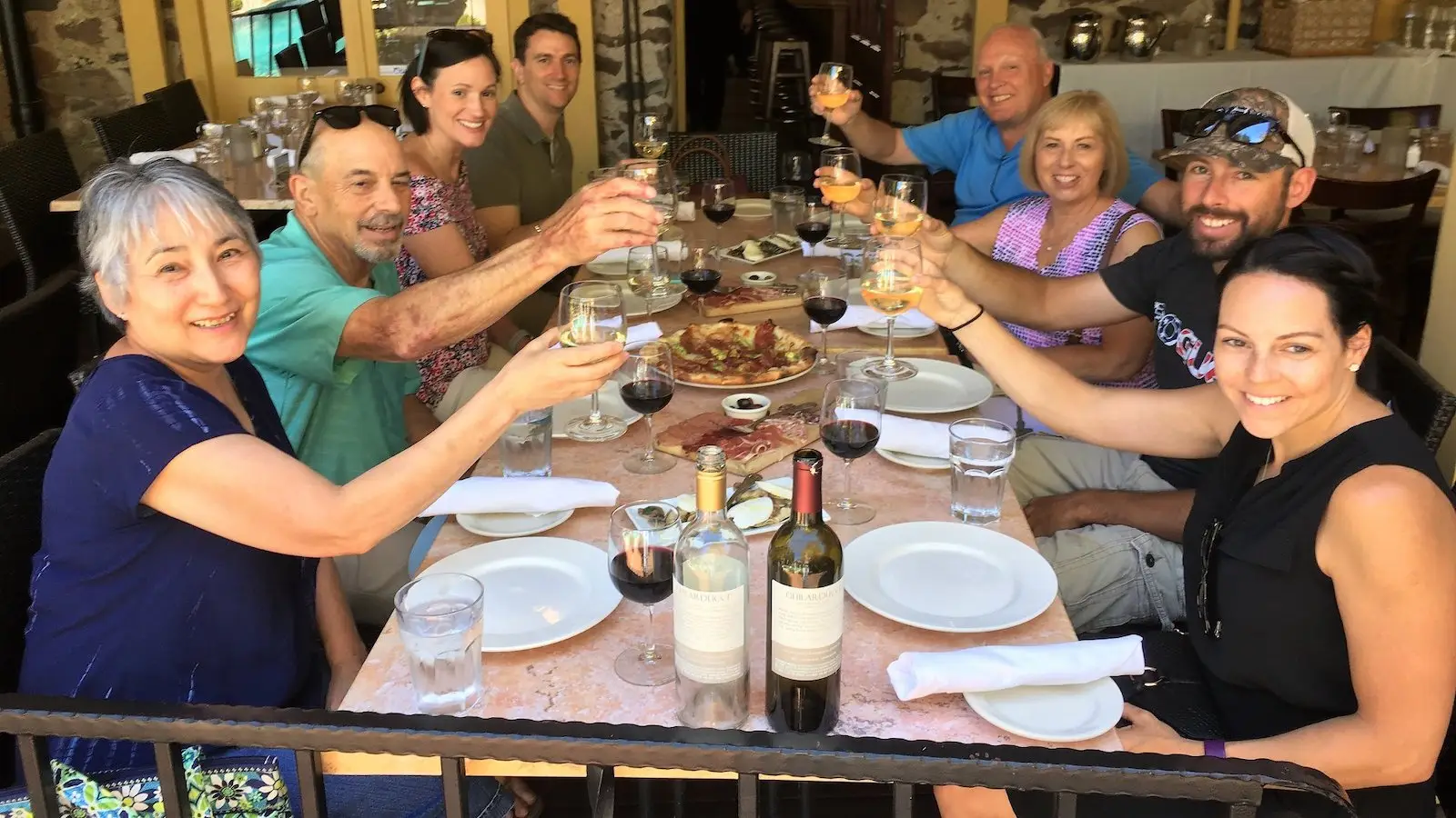 A group of eight people sit around a table outdoors, raising their glasses in a toast. The table is set with plates, wine bottles, water glasses, and a dish of food in the center. The setting appears to be a casual dining restaurant with a festive atmosphere, perfect for one of the top food tours in Sonoma County.