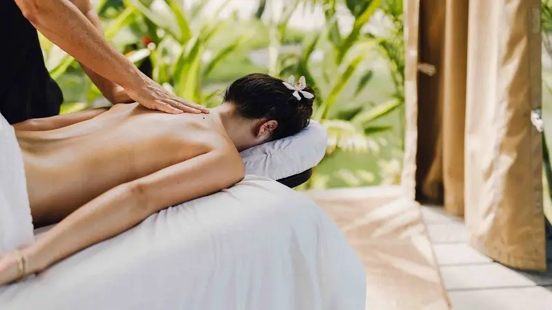 A person lies face down on a massage table outdoors at the best spa hotel in Maui, enjoying a back massage from a therapist. The serene setting features lush green plants and an overall calming atmosphere. A flower in their hair emphasizes the tranquil environment.