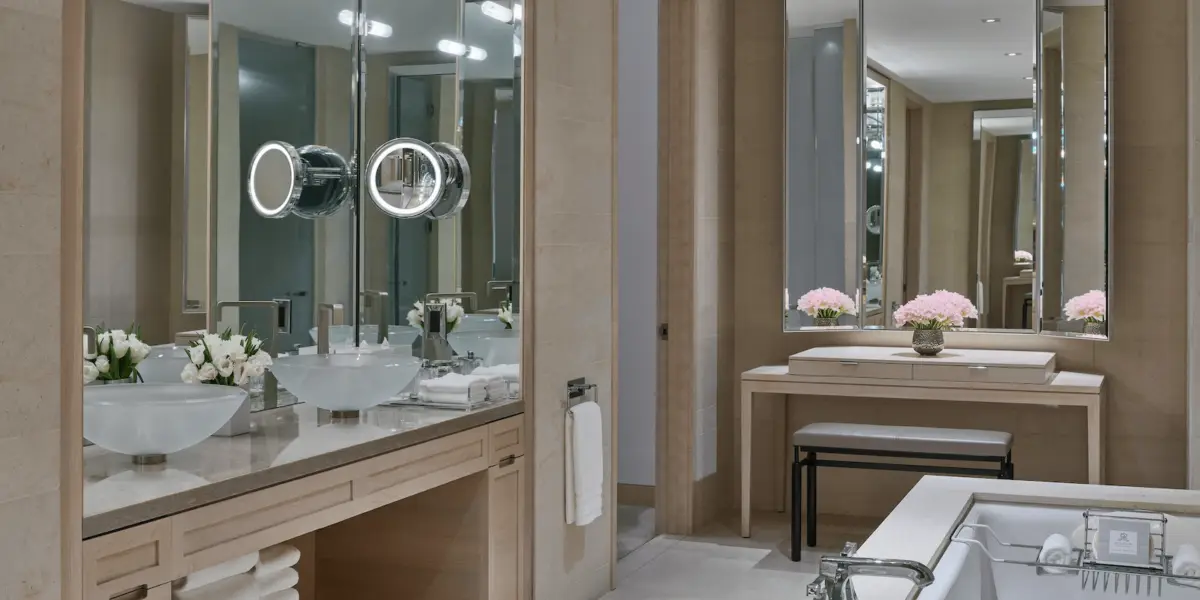 A luxurious bathroom with double sinks, a large mirror with illuminated magnifying mirrors, and fresh flowers. The space features beige tones and modern decor. A vanity area with a small bench and flowers is visible in the background, reminiscent of the best spa hotels San Francisco boasts.