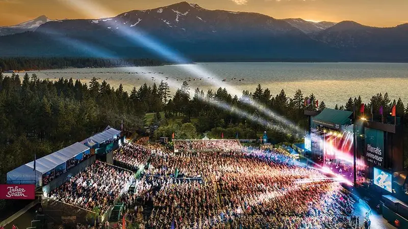 A large outdoor concert at sunset with a packed audience. A stage with bright lights and a screen is on the right, with scenic mountains and a lake in the background. The sky is partly cloudy, and beams of light stretch across the crowd, adding to the vibrant atmosphere—one of the best things to do in Tahoe May.