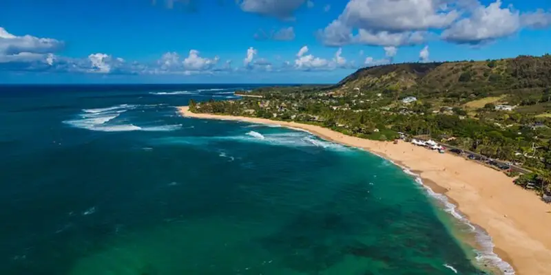 Aerial view of Sunset Beach with turquoise ocean waves gently crashing onto a sandy shore. The shoreline is lined with green vegetation and scattered buildings. Hilly terrain can be seen in the background under a partly cloudy blue sky, capturing the serene essence of this coastal paradise.