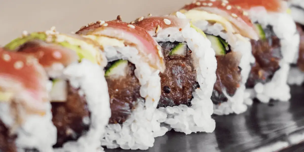 Close-up of the best sushi in North Bay on a dark surface, featuring rice around a central filling of vegetables and possibly meat. Each piece is topped with various ingredients like fish, avocado, and a sprinkling of sesame seeds, showcasing a variety of textures and colors.