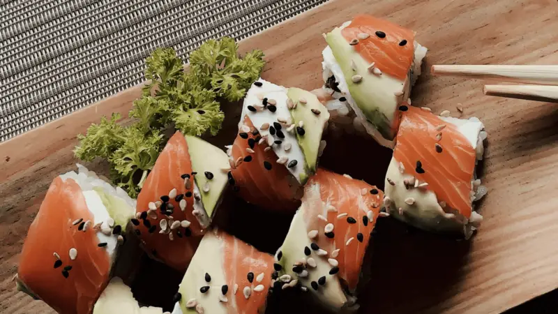 A wooden serving board is topped with sustainable sushi rolls wrapped in salmon and avocado, garnished with black and white sesame seeds. Fresh green parsley rests beside the rolls, and chopsticks are positioned to the right of the sushi. A woven placemat is in the background.