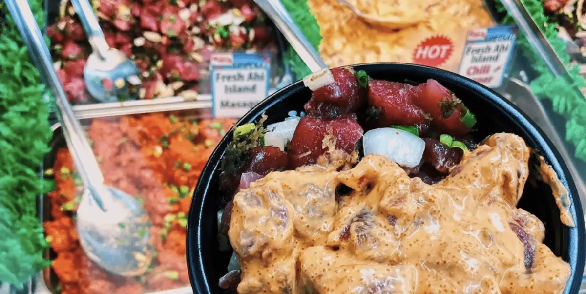 A close-up of a bowl of poke with marinated raw fish, vegetables, and creamy sauce. In the background, there are trays of various poke ingredients and garnishes with visible signs, including one marked "Hot," suggesting a spicy option. This snapshot nods to the rich Poke history interwoven in every ingredient.