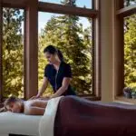 A person lies face down on a massage table, covered partially with a brown blanket, while a massage therapist performs a back massage. Large windows reveal a serene forested view found at some of the best luxury hotels in Tahoe. Massage oils and towels are arranged on a table nearby.