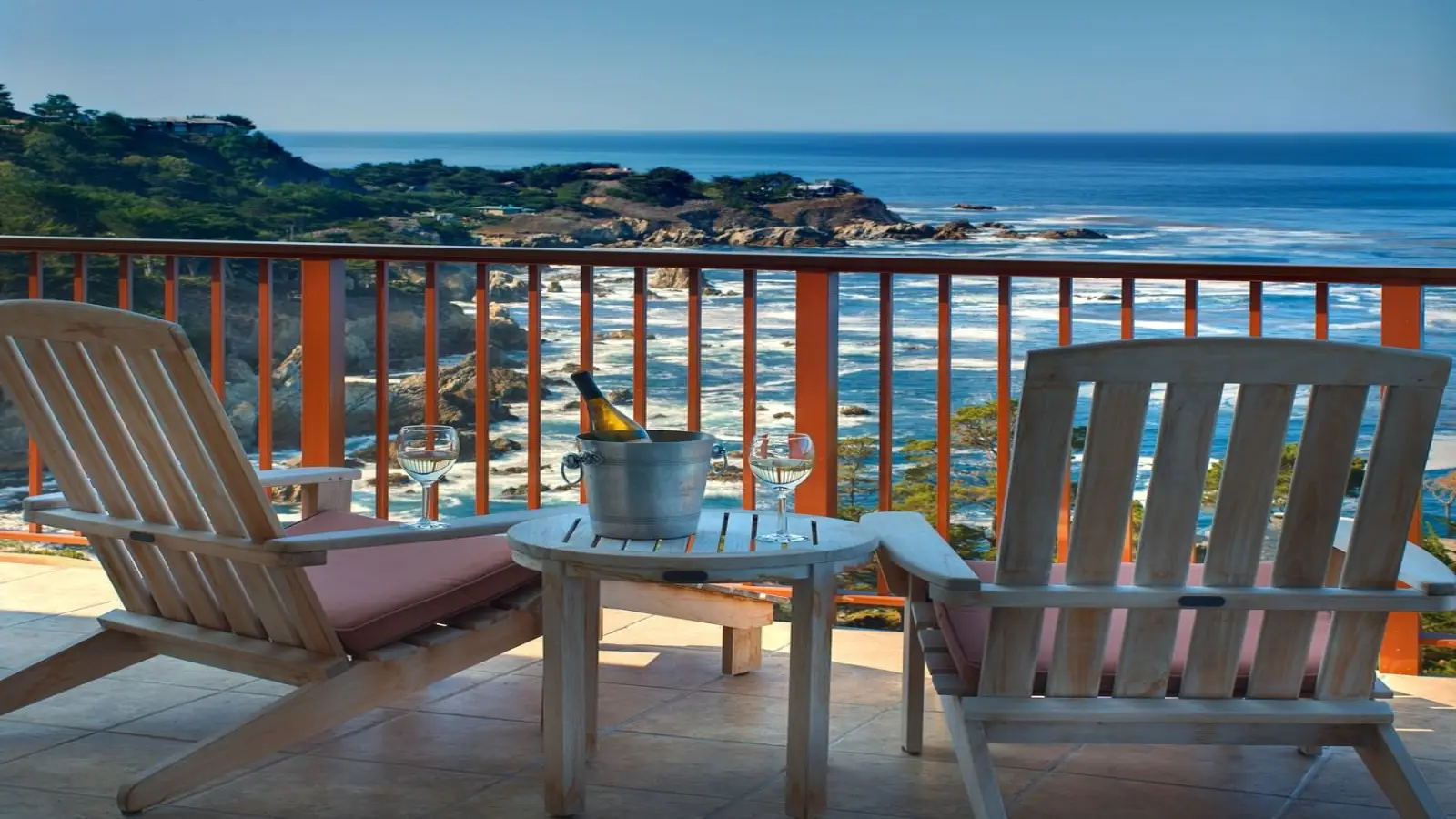 Two wooden chairs with cushions face a scenic ocean view from a balcony in one of the best value hotels on the Monterey Peninsula. A small table between the chairs holds a bucket with champagne and two filled glasses. The coastal landscape features Big Sur’s rocky cliffs and lush greenery under a clear blue sky.