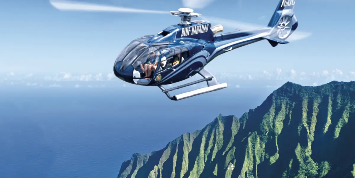 A blue helicopter from "Blue Hawaiian" flies over a scenic coastal landscape with rugged, green mountains and the ocean in the background. The sky is clear with some distant clouds on the horizon—truly one of the best helicopter tours Kauai has to offer.