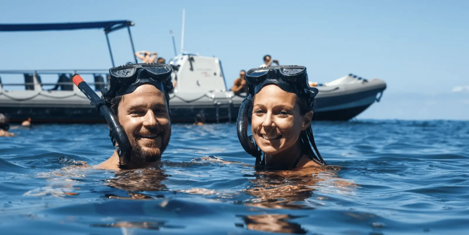 A man and a woman wearing snorkeling masks and snorkels smile while floating in the water. In the background, a boat with several people on board is visible, showcasing one of the best snorkeling tours Kauai has to offer. The sky is clear and blue.