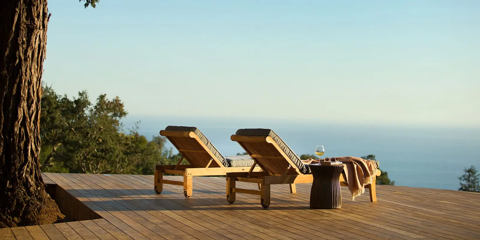 Two wooden lounge chairs are positioned on a wooden deck overlooking a serene ocean view. A side table between the chairs holds a wine glass and a draped blanket. Trees flank the deck, and the scene is bathed in warm sunlight – an idyllic setting reminiscent of the best spa and wellness hotels Monterey Peninsula offers.