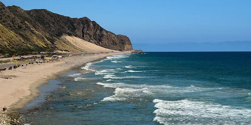 A scenic coastal view with a sandy beach and gentle waves along the shore. A mountainous cliff rises on the left, extending toward the horizon under a clear blue sky. The beach stretches alongside a road with small structures and parked vehicles, capturing the charm of a Four-Days Ventura County Coast getaway.