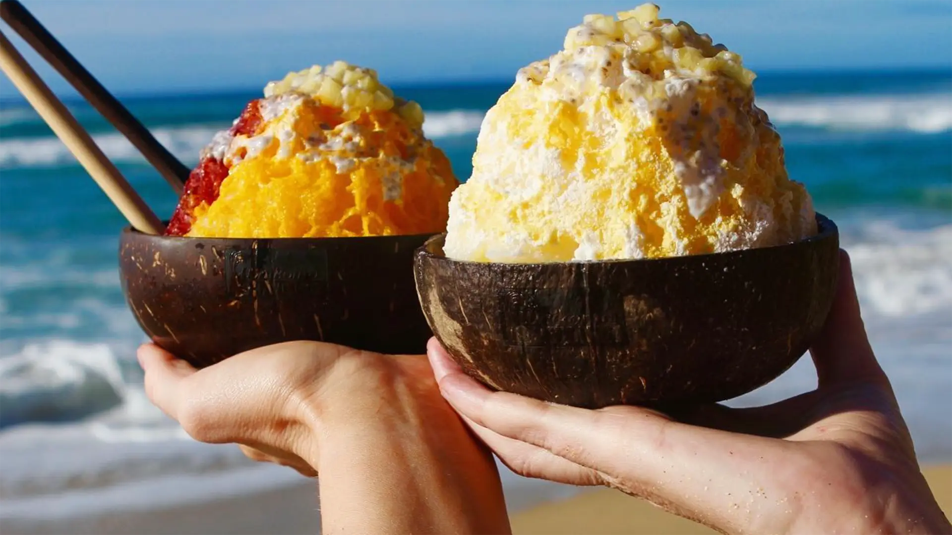 Two hands holding wooden bowls of the best shave ice Kauai offers, colorful desserts topped with various fruits. One is yellow and red, while the other is predominantly yellow. In the background, a sandy beach and ocean waves under a clear blue sky complete this idyllic scene.