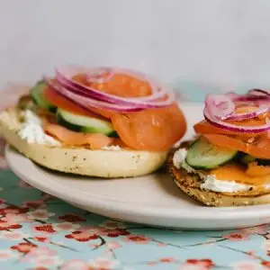 A beige plate holds an open-faced bagel topped with cream cheese, smoked salmon, cucumber slices, and red onion rings. The background features a patterned tablecloth with floral designs—perfect for those wondering where to eat in Tahoe.