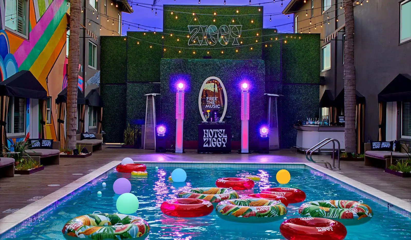 A vibrant pool area at Hotel Ziggy is decorated with colorful inflatable rings and glowing orbs floating in the water. The backdrop features lush greenery, string lights, and a DJ booth with "FREE THE MUSIC" signage, creating a lively, festive atmosphere at Hotel Ziggy.