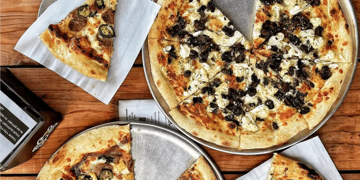 Three metal plates hold different pizzas, each with a slice removed. The best pizza in the East Bay is placed on a wooden surface, topped with olives, mushrooms, and cheese. A container with paper napkins is visible on the left side of the image.