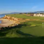 A coastal golf course features a green next to a sandy beach, with waves crashing on the rocky shore. A few people are walking along the beach, and several houses are situated in the distance. The backdrop includes mountains under a clear blue sky—one of the best golf courses in the greater bay area.