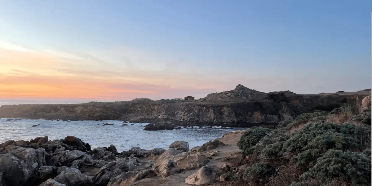 Rocky seaside landscape during sunset. The view shows a rugged coastline with rocks and cliffs, sparse green vegetation on the right, and calm waves washing against the shore. The sky is painted with hues of orange and pink, gradually blending into a clear blue, perfect for the best scuba diving Northern California offers.